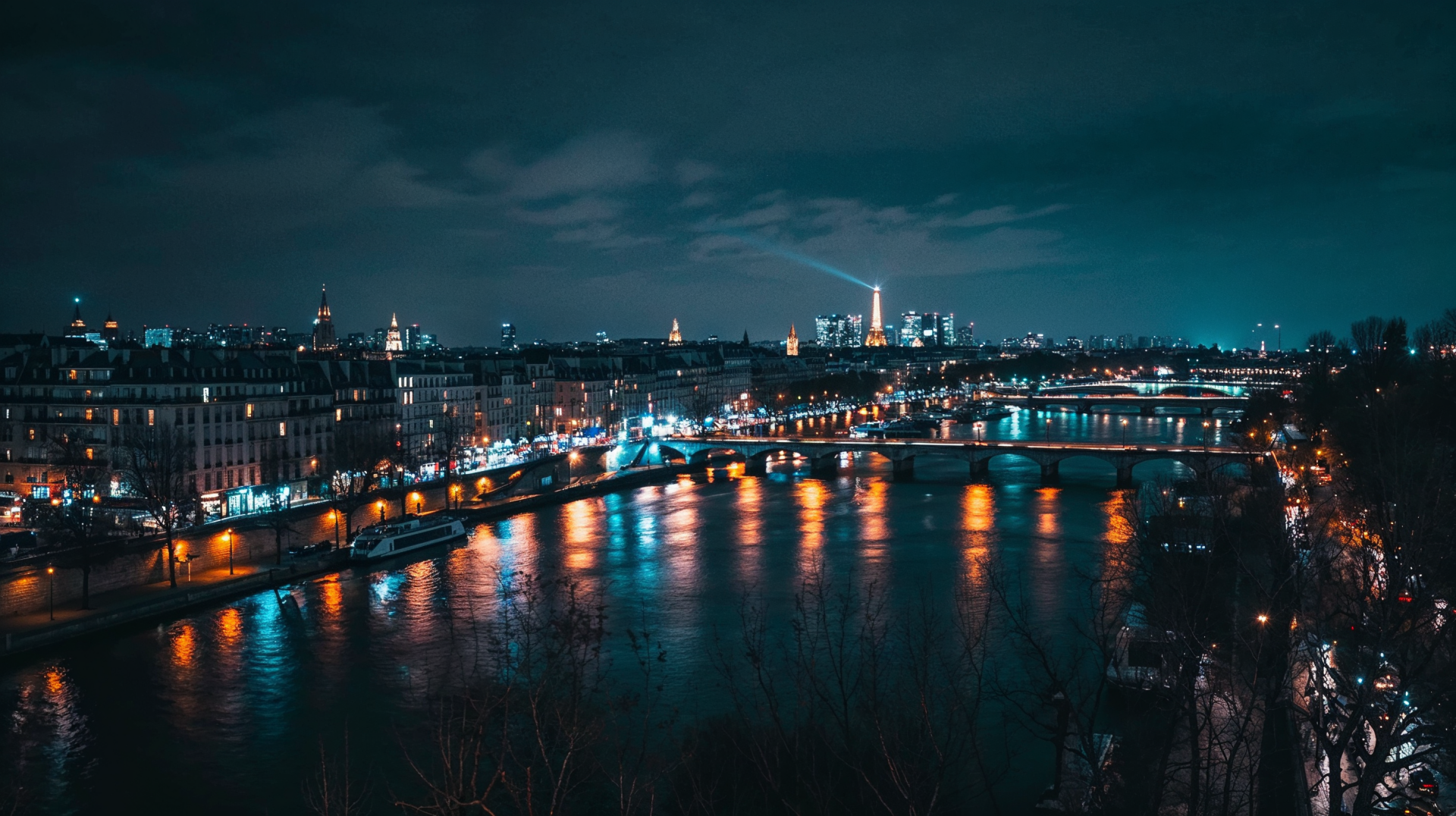 HD PC desktop wallpaper of Paris at night, showcasing the illuminated cityscape and bridges reflecting on the Seine River under a moody, cloudy sky.