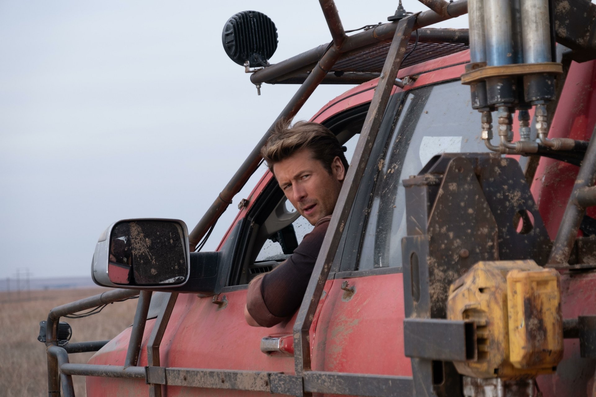 HD desktop wallpaper featuring Glen Powell from the movie Twisters, leaning out of a rugged, red vehicle in an open field.