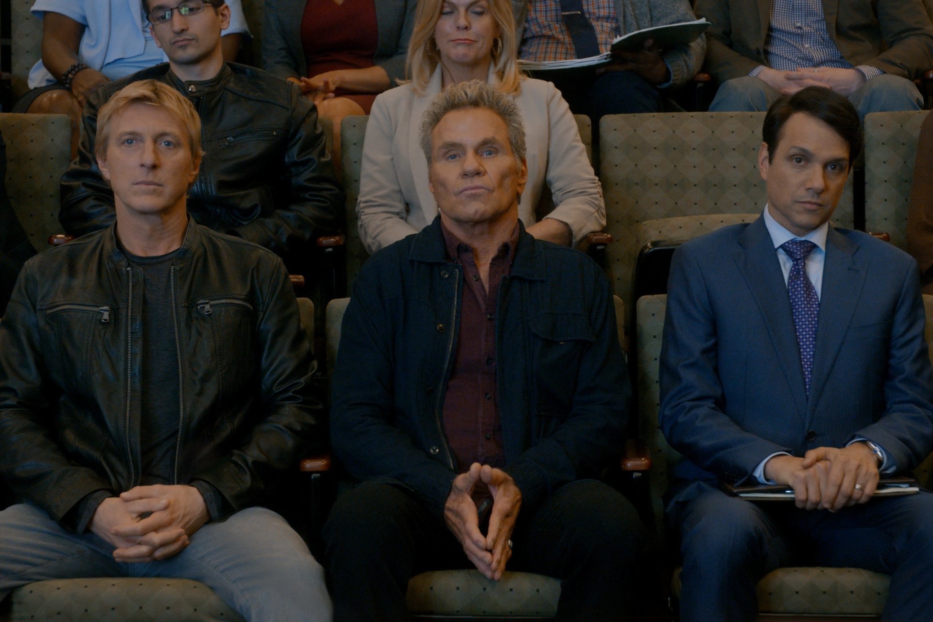 HD desktop wallpaper featuring characters from the TV show Cobra Kai seated in an auditorium, portraying a tense moment.