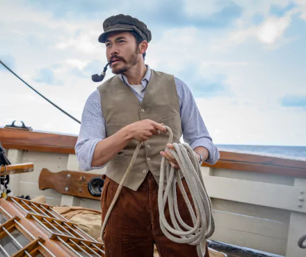HD desktop wallpaper depicting a scene from The Ministry of Ungentlemanly Warfare featuring a man dressed in historical attire with a waistcoat and a cap, standing on the deck of a ship, holding a rope.