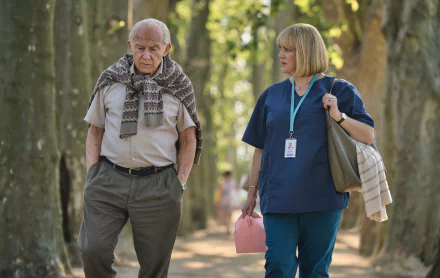 HD desktop wallpaper from The Tattooist of Auschwitz TV show featuring an elderly man and a woman in scrubs walking together under tree-lined pathway.