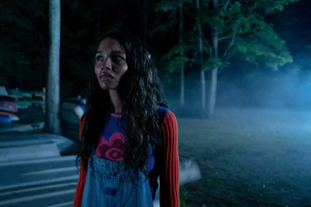 HD desktop wallpaper from the movie I Saw the TV Glow featuring a young woman standing anxiously in a dimly lit, foggy forest at night.