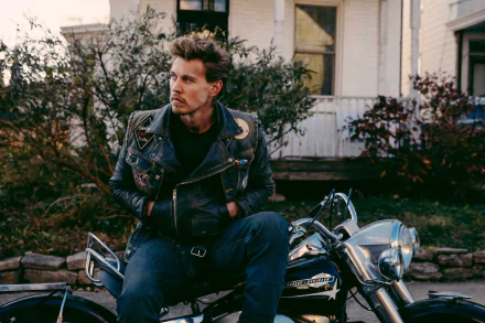 HD desktop wallpaper featuring a scene from the movie The Bikeriders, with Austin Butler sitting on a motorcycle, wearing a leather jacket, in front of a rustic house and greenery in the background.