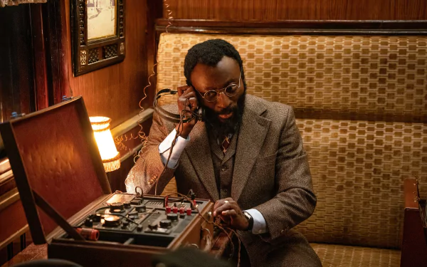 A man dressed in a vintage suit sits in an ornate room, working attentively on a complex device inside a briefcase. This image is a HD desktop wallpaper related to the movie The Ministry of Ungentlemanly Warfare.
