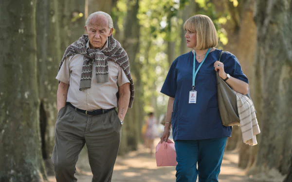 HD desktop wallpaper from The Tattooist of Auschwitz TV show featuring an elderly man and a woman in scrubs walking together under tree-lined pathway.