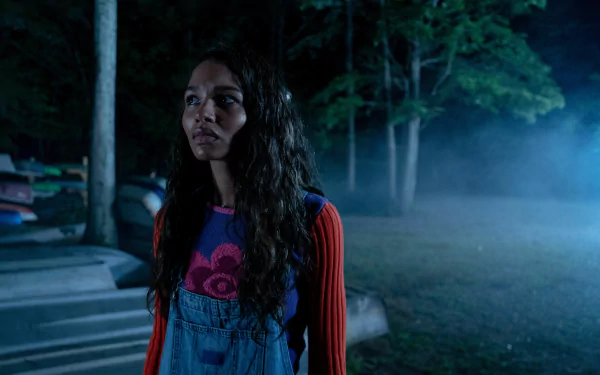 HD desktop wallpaper from the movie I Saw the TV Glow featuring a young woman standing anxiously in a dimly lit, foggy forest at night.