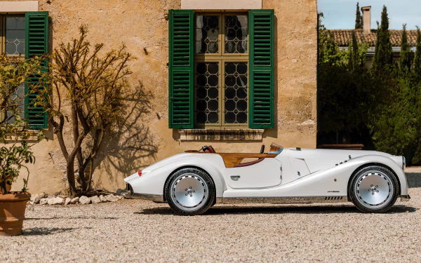 HD desktop wallpaper of a classic white Morgan Midsummer car parked in front of a rustic building with green shutters, set against a serene outdoor background.