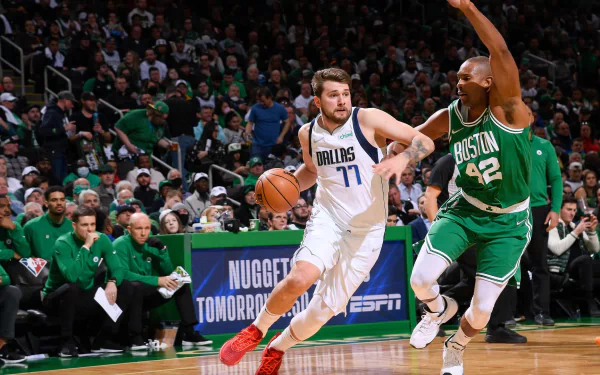 Luka Dončić of the Dallas Mavericks dribbles past a Boston Celtics defender during an NBA game. The crowded arena captures the intense atmosphere, making this a captivating HD desktop wallpaper and background.