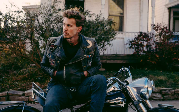 HD desktop wallpaper featuring a scene from the movie The Bikeriders, with Austin Butler sitting on a motorcycle, wearing a leather jacket, in front of a rustic house and greenery in the background.