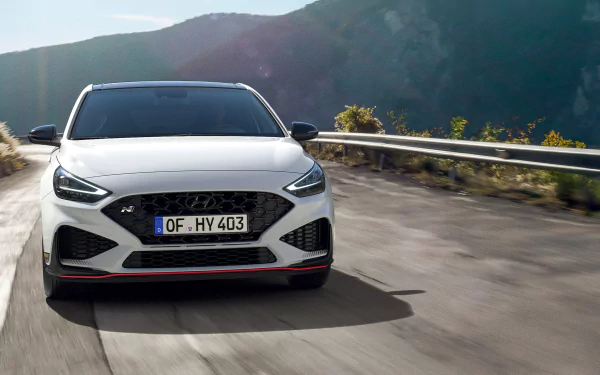 HD wallpaper of a Hyundai i30 car driving on a mountain road, captured from the front with scenic mountainous background. The image emphasizes the sleek design of the vehicle.