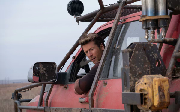 HD desktop wallpaper featuring Glen Powell from the movie Twisters, leaning out of a rugged, red vehicle in an open field.