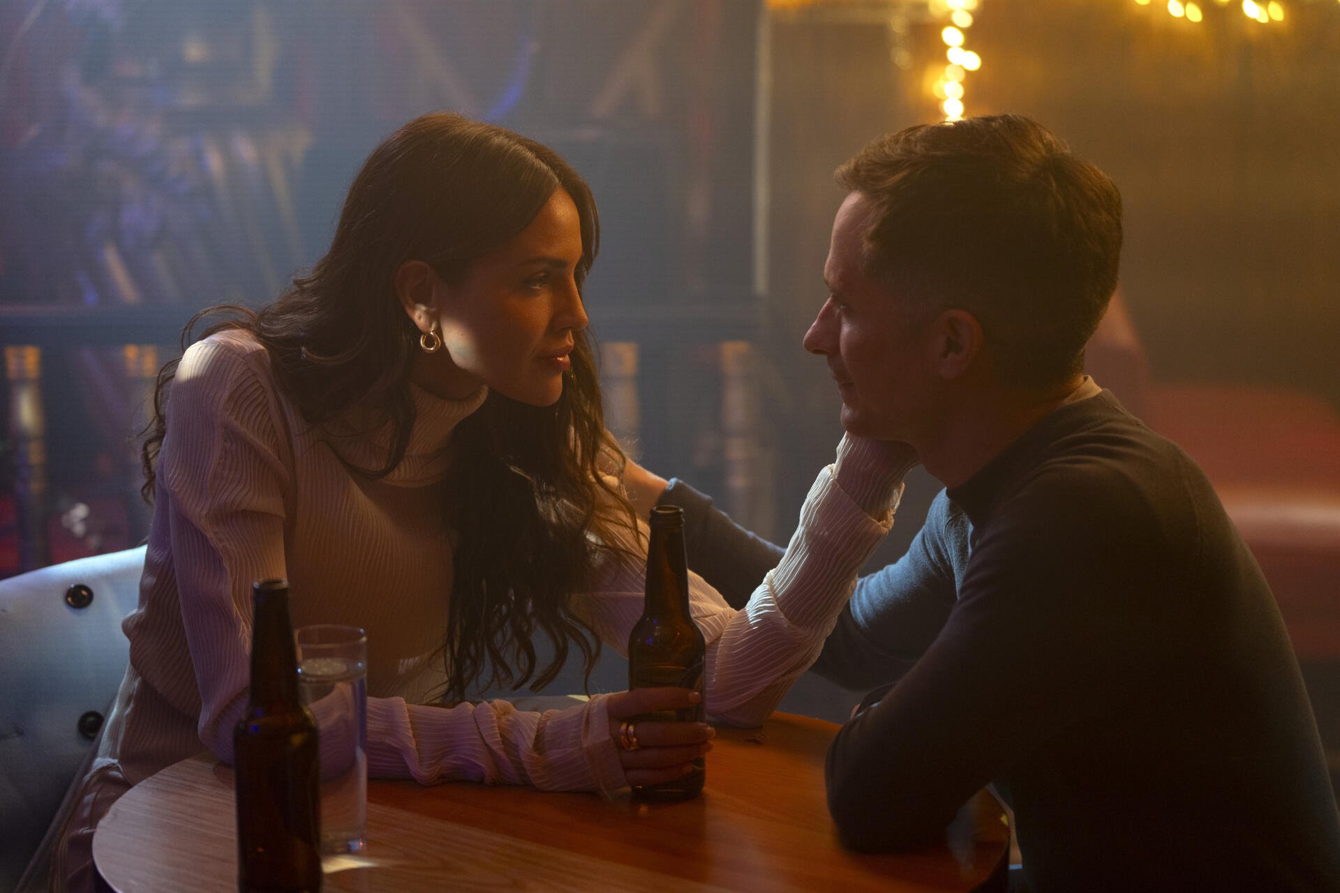 A scene from the TV show La Máquina (2024), featuring two characters in an intimate conversation at a bar, with warm lighting and drinks on the table.