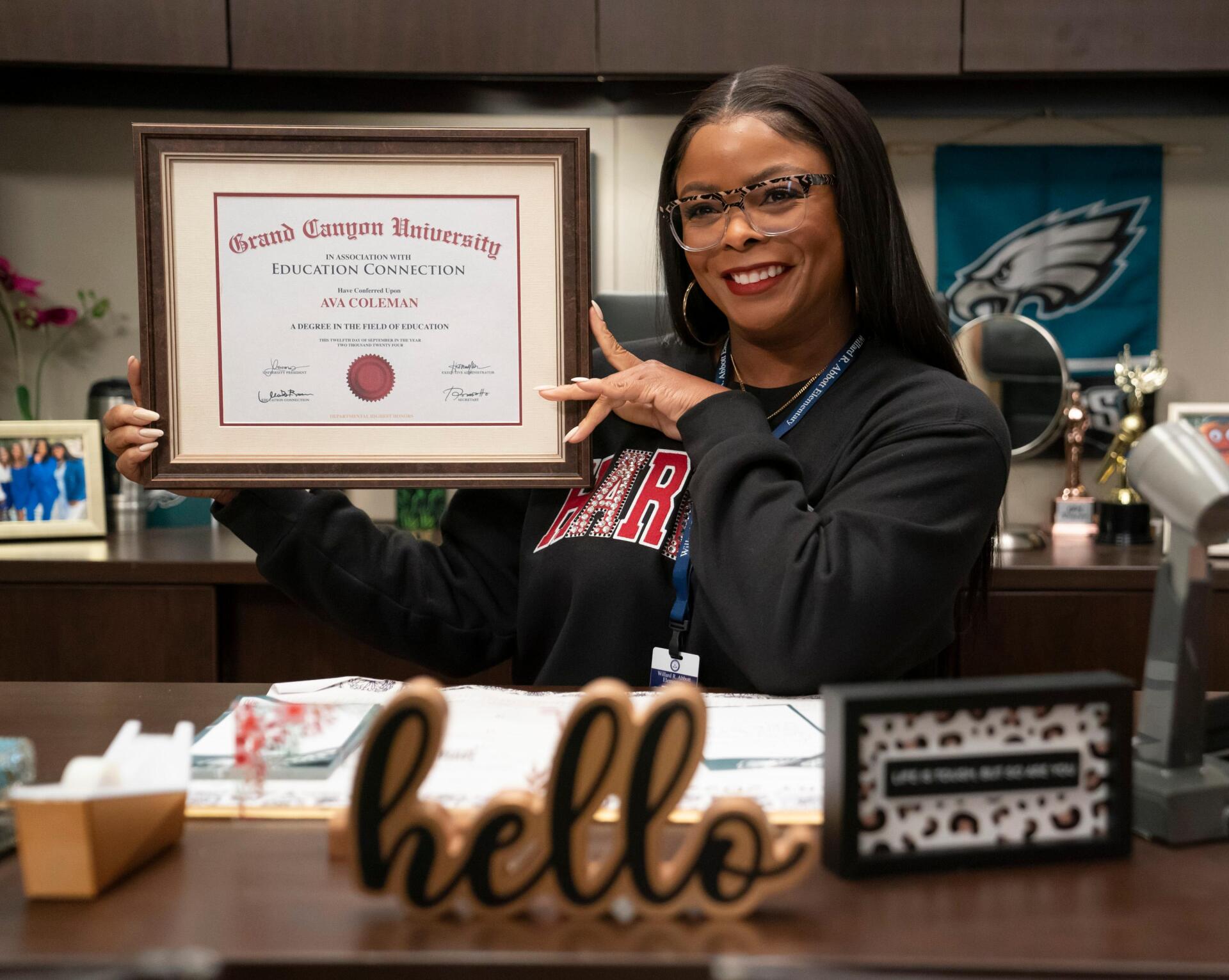 A character from the TV show Abbott Elementary proudly holds up a diploma from Grand Canyon University, smiling in an office setting adorned with personal touches.