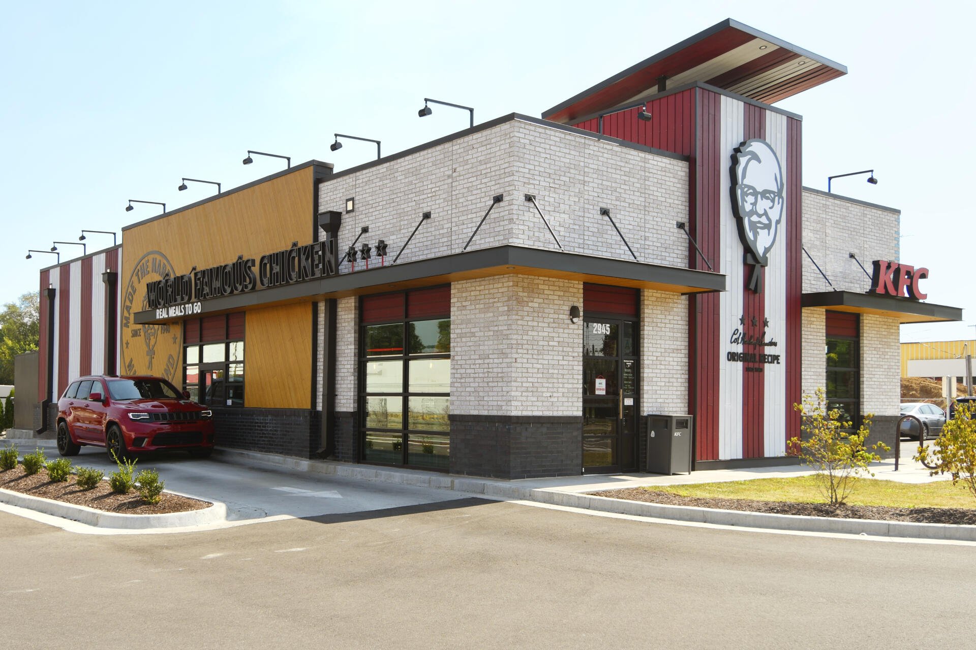 A modern KFC restaurant featuring a stylish exterior with a blend of brick and bold colors, surrounded by landscaping and a parking area, serving as an HD desktop wallpaper.