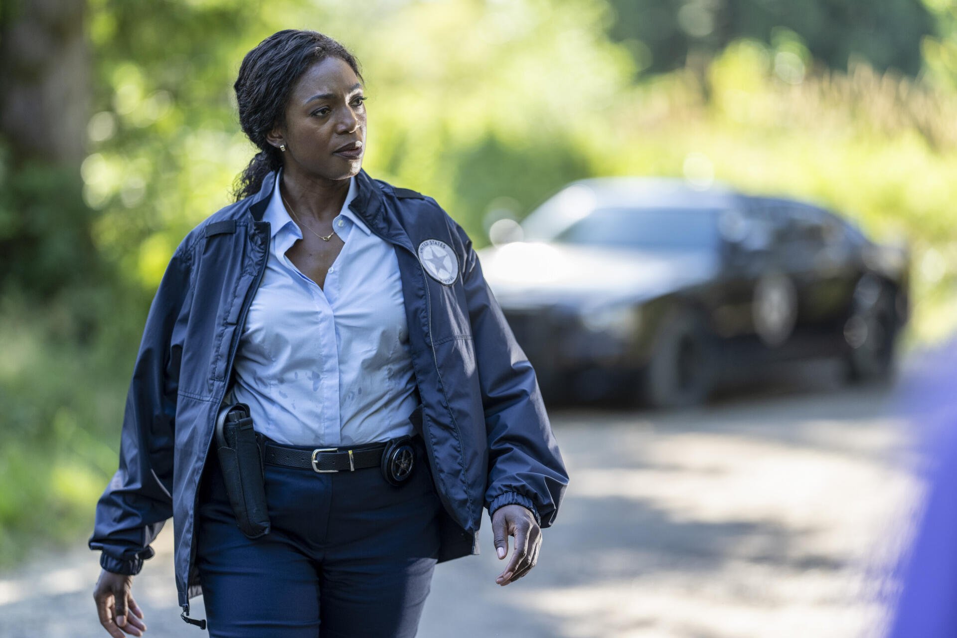 A scene from the TV show *Tracker* (2024), featuring a woman in a uniform walking along a road, with a police car in the background, set in a natural landscape.