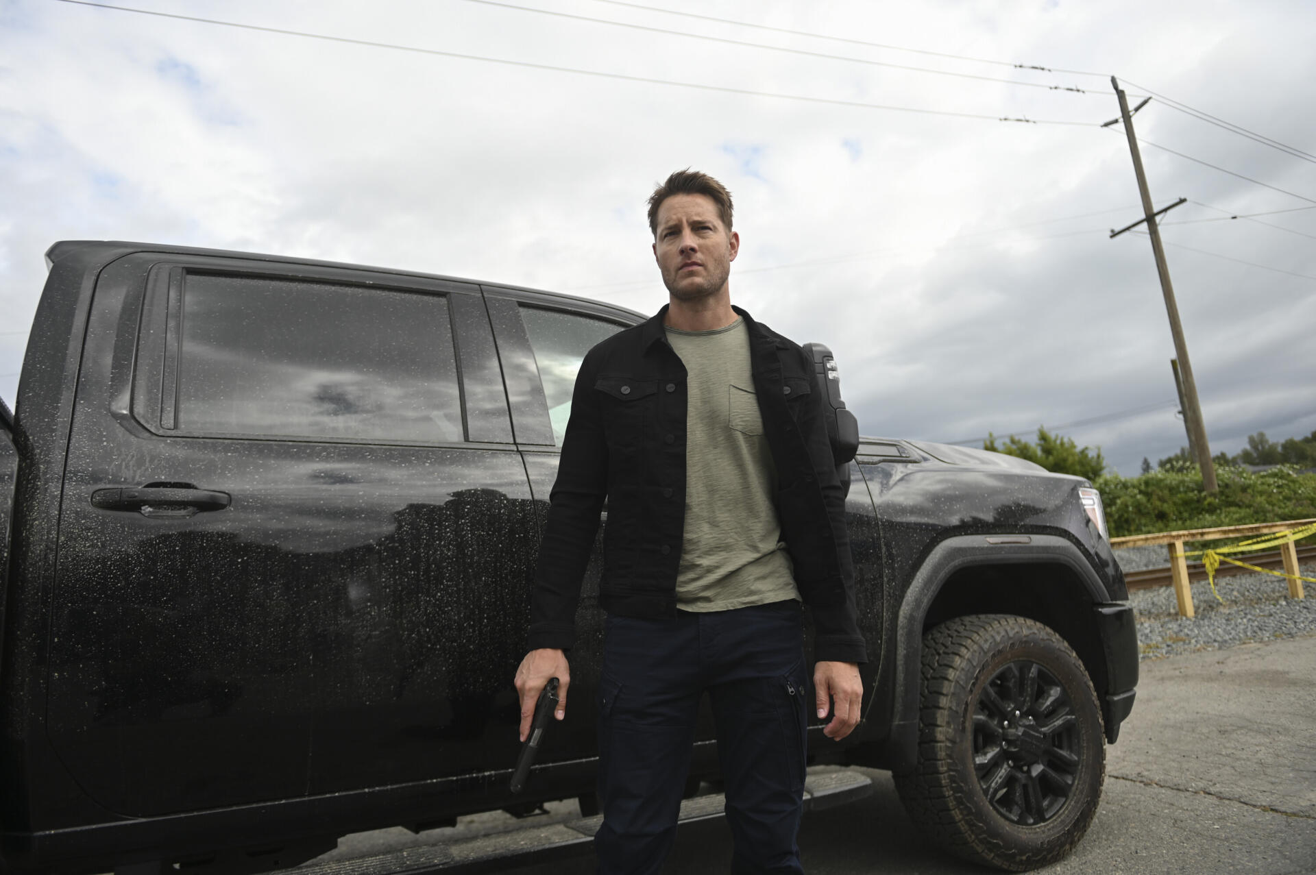 A HD desktop wallpaper featuring Justin Hartley from the TV show Tracker (2024), standing confidently beside a black truck in an atmospheric outdoor setting.