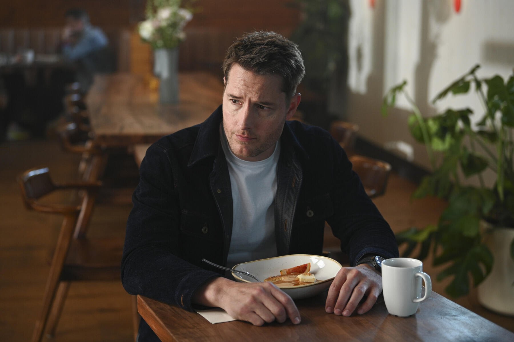 Justin Hartley appears contemplative at a dining table in a scene from the TV show Tracker (2024), featuring a warm, inviting background with plants and soft lighting.
