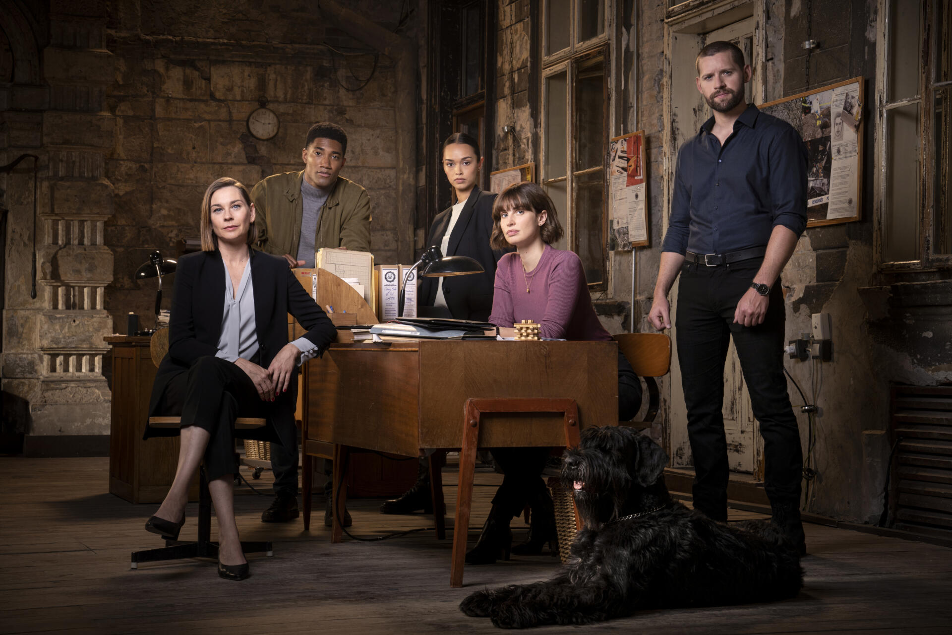 A group of five characters from the TV show FBI: International stands in a dimly lit room, with one seated at a desk and a dog nearby, creating an engaging desktop wallpaper.