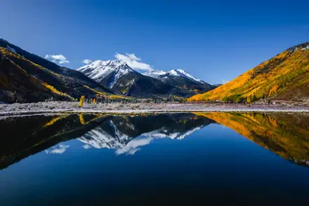 A serene landscape of Crystal Lake reflecting majestic mountains under a clear blue sky, framed by vibrant autumn foliage. This stunning 4K Ultra HD wallpaper captures nature's beauty.