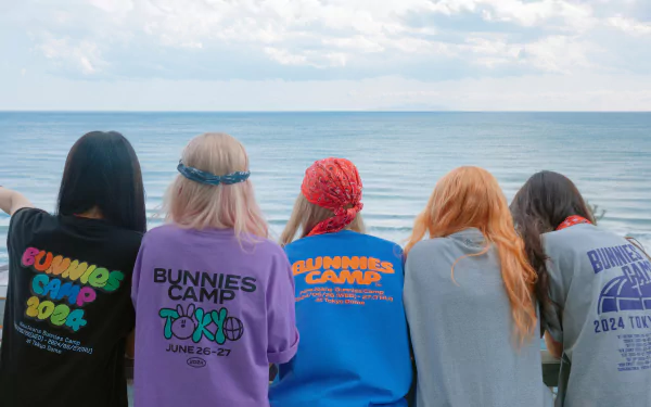 A vibrant HD wallpaper featuring five friends in colorful sweatshirts, enjoying the ocean view, capturing a carefree moment tied to NewJeans music culture.