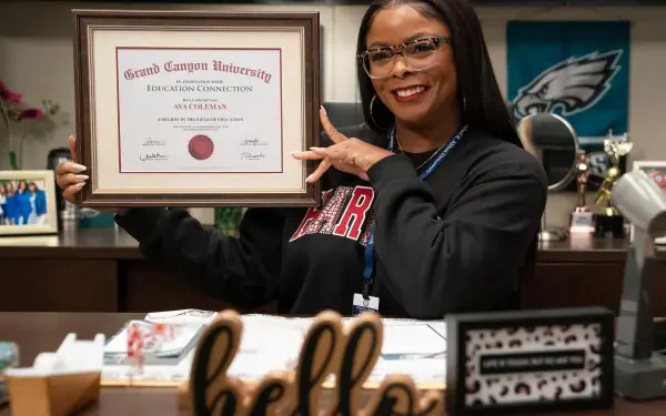 A character from the TV show Abbott Elementary proudly holds up a diploma from Grand Canyon University, smiling in an office setting adorned with personal touches.