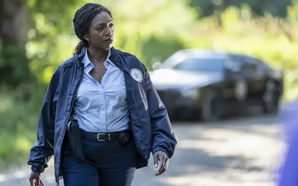 A scene from the TV show *Tracker* (2024), featuring a woman in a uniform walking along a road, with a police car in the background, set in a natural landscape.