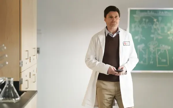 A scene from the TV show Tracker (2024), featuring a person in a lab coat standing in a classroom with a chalkboard, showcasing a blend of professionalism and intrigue.