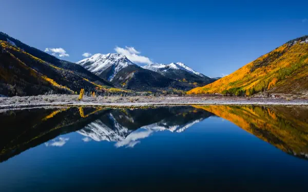 A serene landscape of Crystal Lake reflecting majestic mountains under a clear blue sky, framed by vibrant autumn foliage. This stunning 4K Ultra HD wallpaper captures nature's beauty.