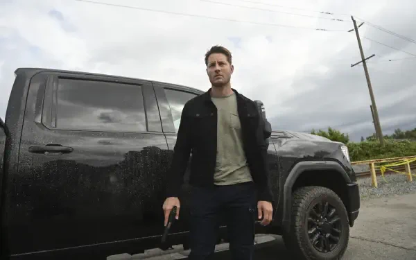 A HD desktop wallpaper featuring Justin Hartley from the TV show Tracker (2024), standing confidently beside a black truck in an atmospheric outdoor setting.
