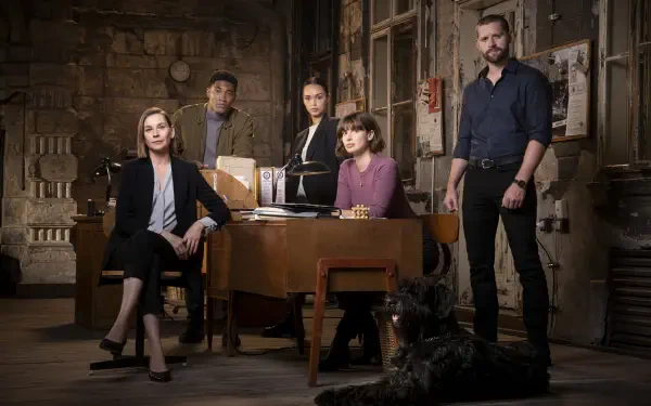 A group of five characters from the TV show FBI: International stands in a dimly lit room, with one seated at a desk and a dog nearby, creating an engaging desktop wallpaper.