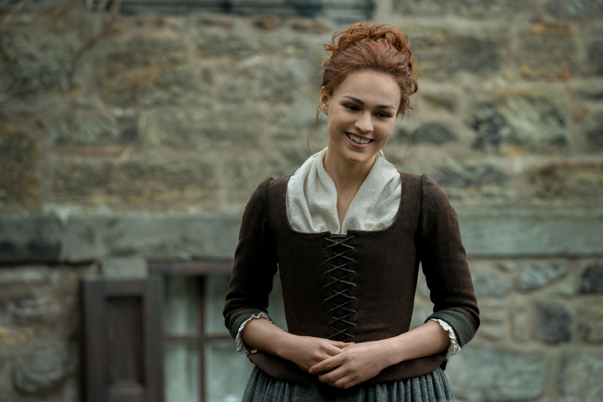 Sophie Skelton smiles in a period costume against a stone backdrop, capturing the essence of her character in the TV show Outlander. A stunning 4K Ultra HD desktop wallpaper.