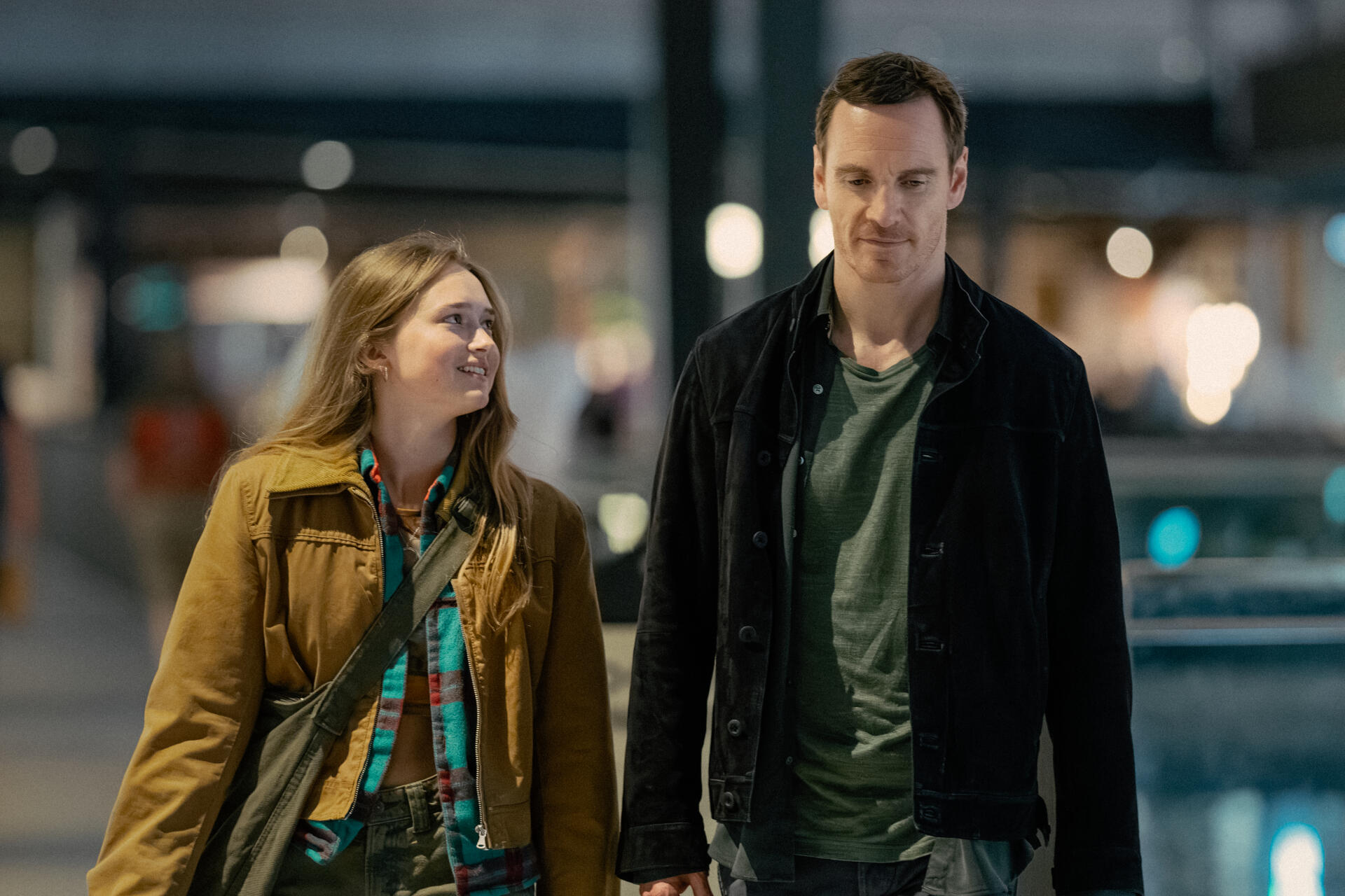 Two characters walk together in a modern setting, smiling and engaging in conversation. This image is a scene from the TV show The Agency, featuring Michael Fassbender.