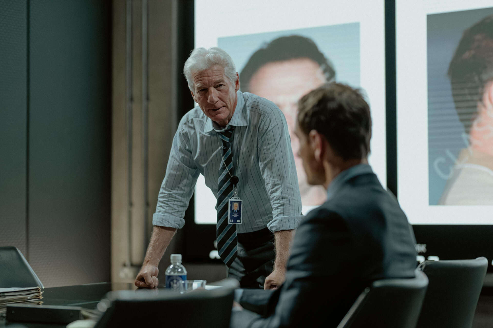 A scene from the TV show The Agency featuring Richard Gere, showcasing a tense discussion in a modern meeting room with images projected on a screen.