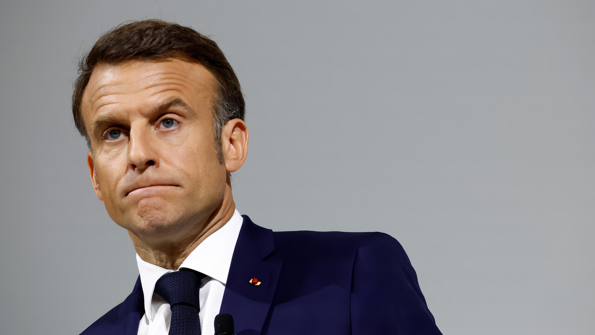 Emmanuel Macron, dressed in a suit and tie, is captured in a thoughtful pose against a neutral background, creating a striking HD desktop wallpaper and background.