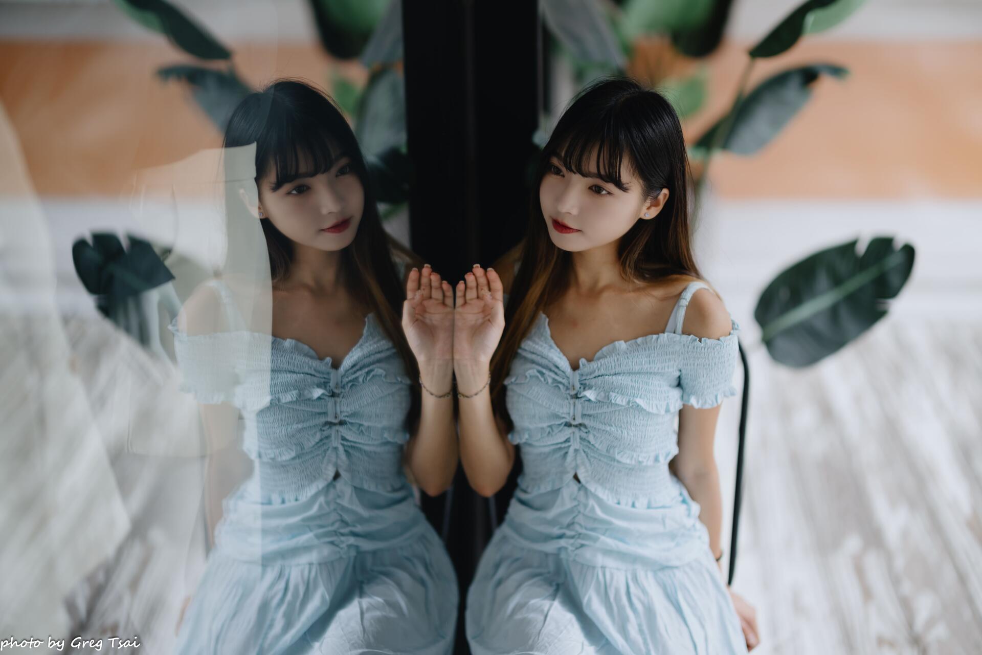 Woman in a pale blue dress touching a glass reflection against a soft leafy backdrop — 8K Ultra HD PC desktop wallpaper.