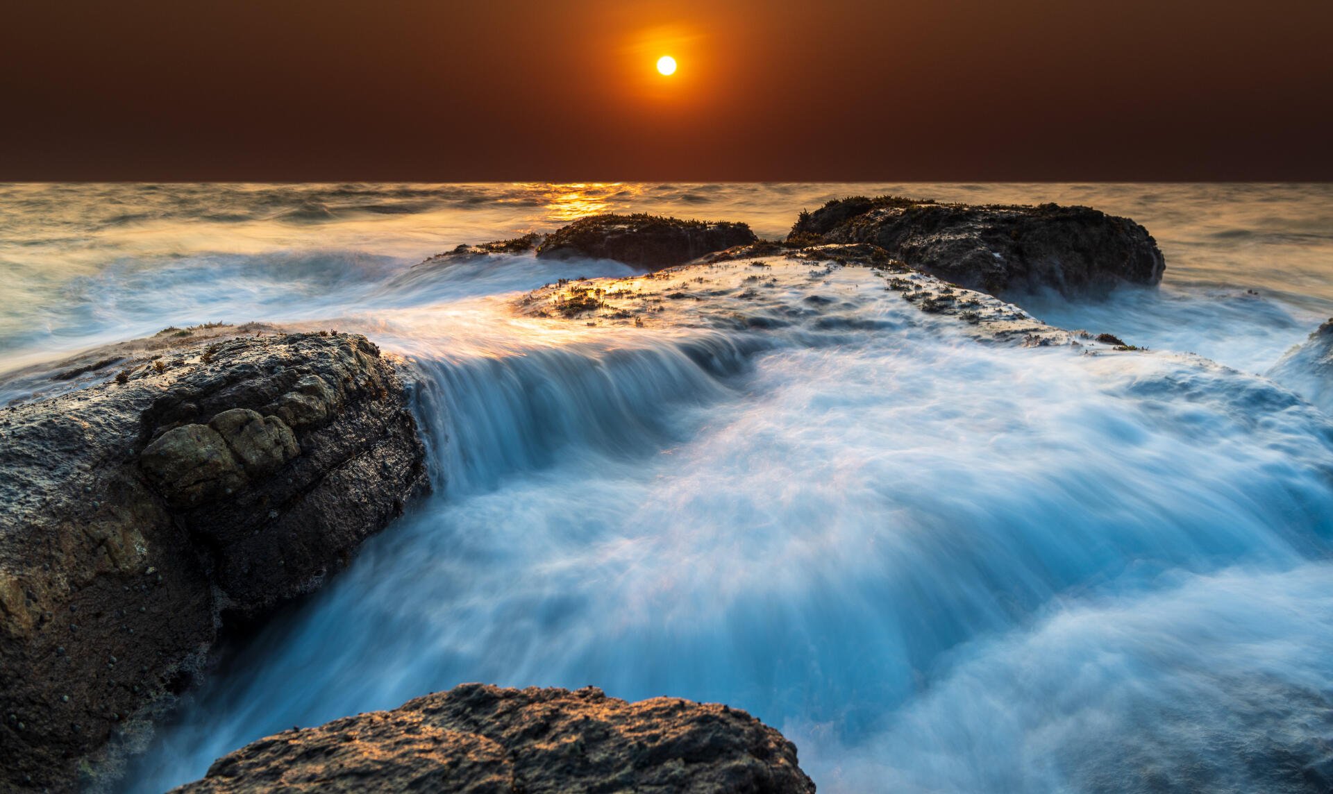 5K Ultra HD PC desktop wallpaper: landscape water scene — coastal sunset with an orange sun over the horizon, waves cascading over rocky shore with silky blue-white surf.