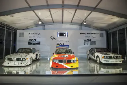 An HD wallpaper featuring three iconic BMW models: the 3.0 CSL, 635CSi, and a vintage racing variant, displayed in a modern exhibition setting under bright lights.