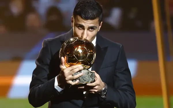 Rodri Hernández, dressed in a sharp suit, kisses the Ballon d'Or while celebrating his achievement in soccer, showcasing his triumph in the sports world. HD desktop wallpaper background.