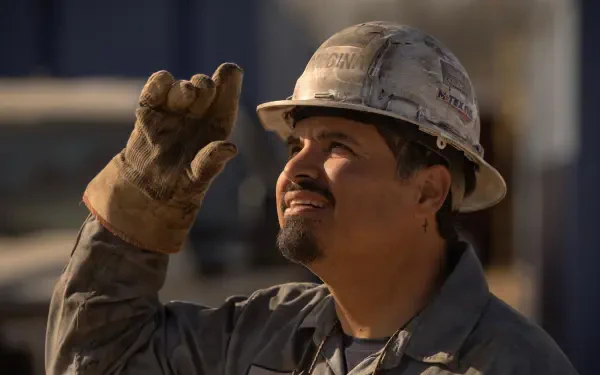 A still from the TV show Landman, featuring Michael Peña in a hard hat and work gloves, looking up with a smile, set against a construction background. 4K Ultra HD wallpaper.