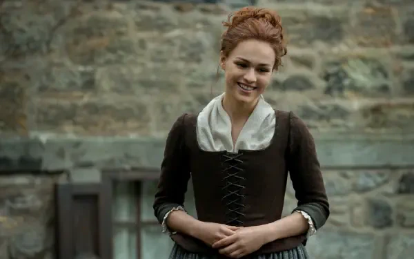 Sophie Skelton smiles in a period costume against a stone backdrop, capturing the essence of her character in the TV show Outlander. A stunning 4K Ultra HD desktop wallpaper.