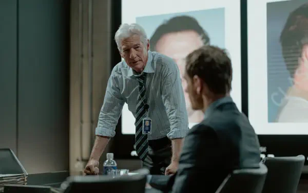 A scene from the TV show The Agency featuring Richard Gere, showcasing a tense discussion in a modern meeting room with images projected on a screen.