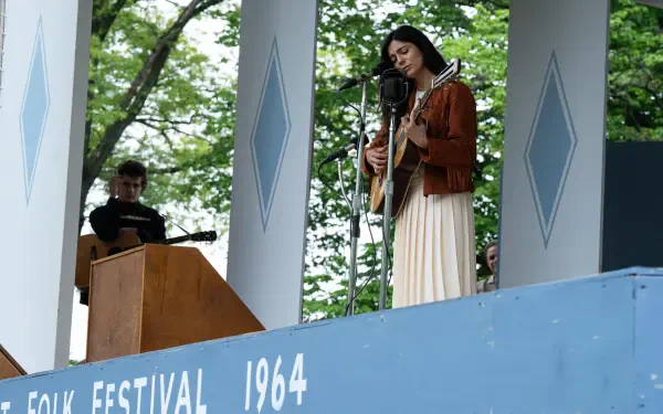 A woman performs as a singer on stage at a 1964 folk festival, featured in the movie A Complete Unknown, showcasing Monica Barbaro in an 8K Ultra HD scene.