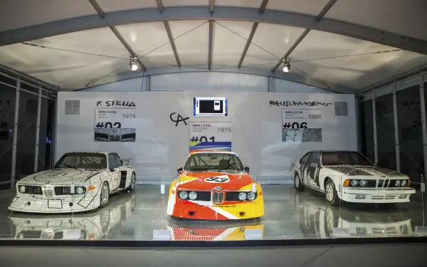 An HD wallpaper featuring three iconic BMW models: the 3.0 CSL, 635CSi, and a vintage racing variant, displayed in a modern exhibition setting under bright lights.