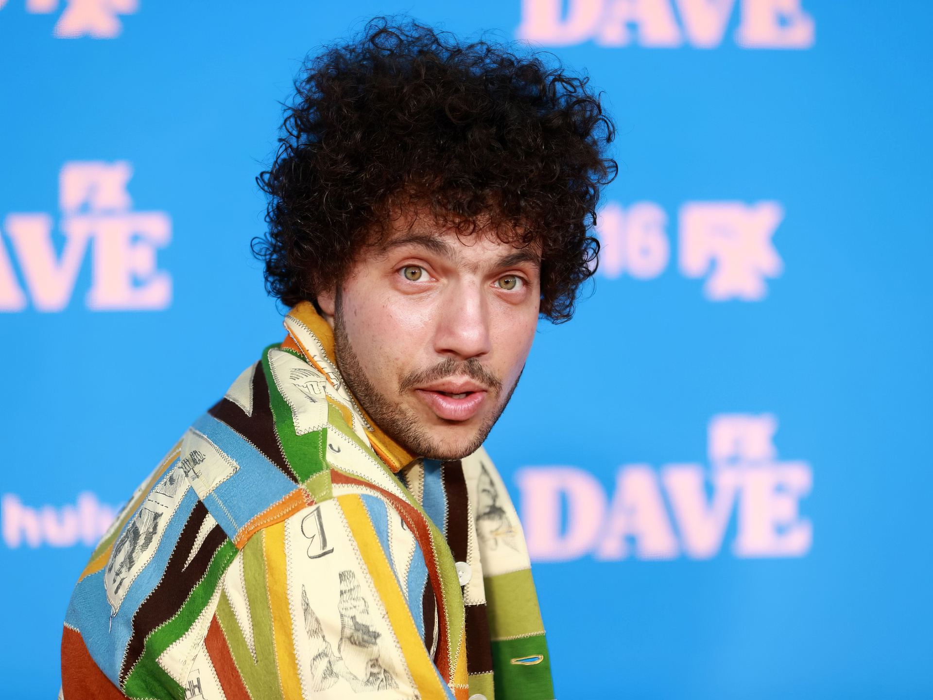 A vibrant HD desktop wallpaper featuring Benny Blanco, showcasing his distinctive fashion and engaging expression against a bright blue background.