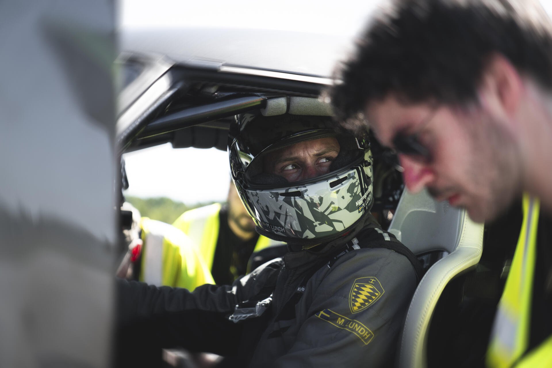 A car racer in a helmet sits in a sleek vehicle, engaging with a teammate in sunglasses. An 8K Ultra HD wallpaper showcasing the thrill of motorsport.