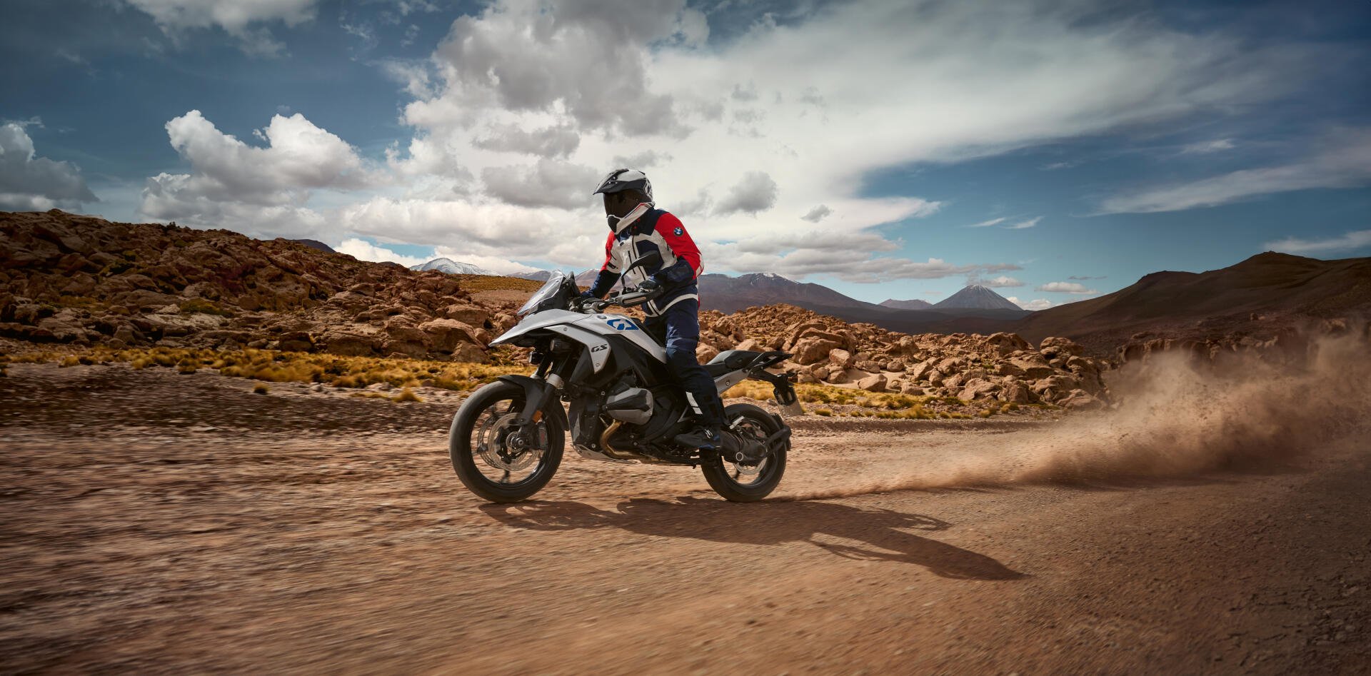 A BMW R1300GS motorcycle navigates a rugged dirt path, capturing a thrilling moment in motocross against a backdrop of dramatic clouds and mountains. 4K Ultra HD quality.