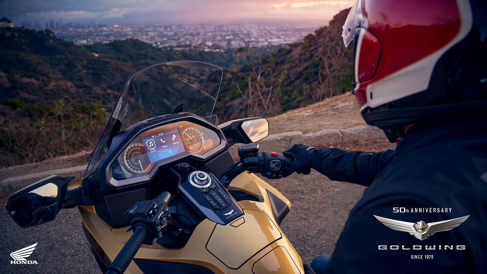 A Honda GL 1800 Gold Wing motorcycle parked on a scenic overlook, with a rider in a red helmet gazing at a vibrant sunset over a cityscape in the background.