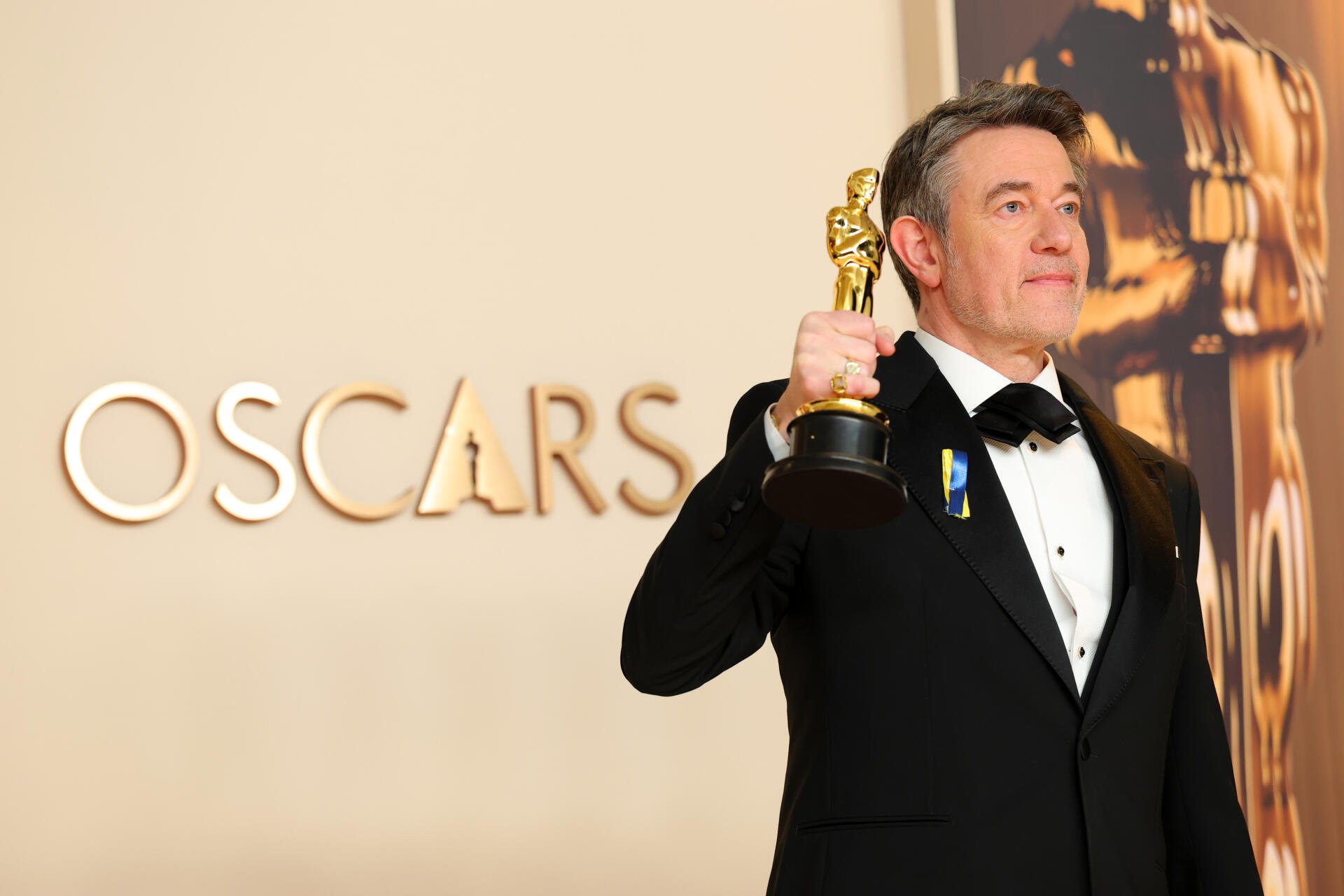 Peter Straughan proudly holds an Oscar trophy, dressed in formal attire, with the iconic OSCARS backdrop. This 4K Ultra HD image captures a moment of celebration at the Academy Awards.