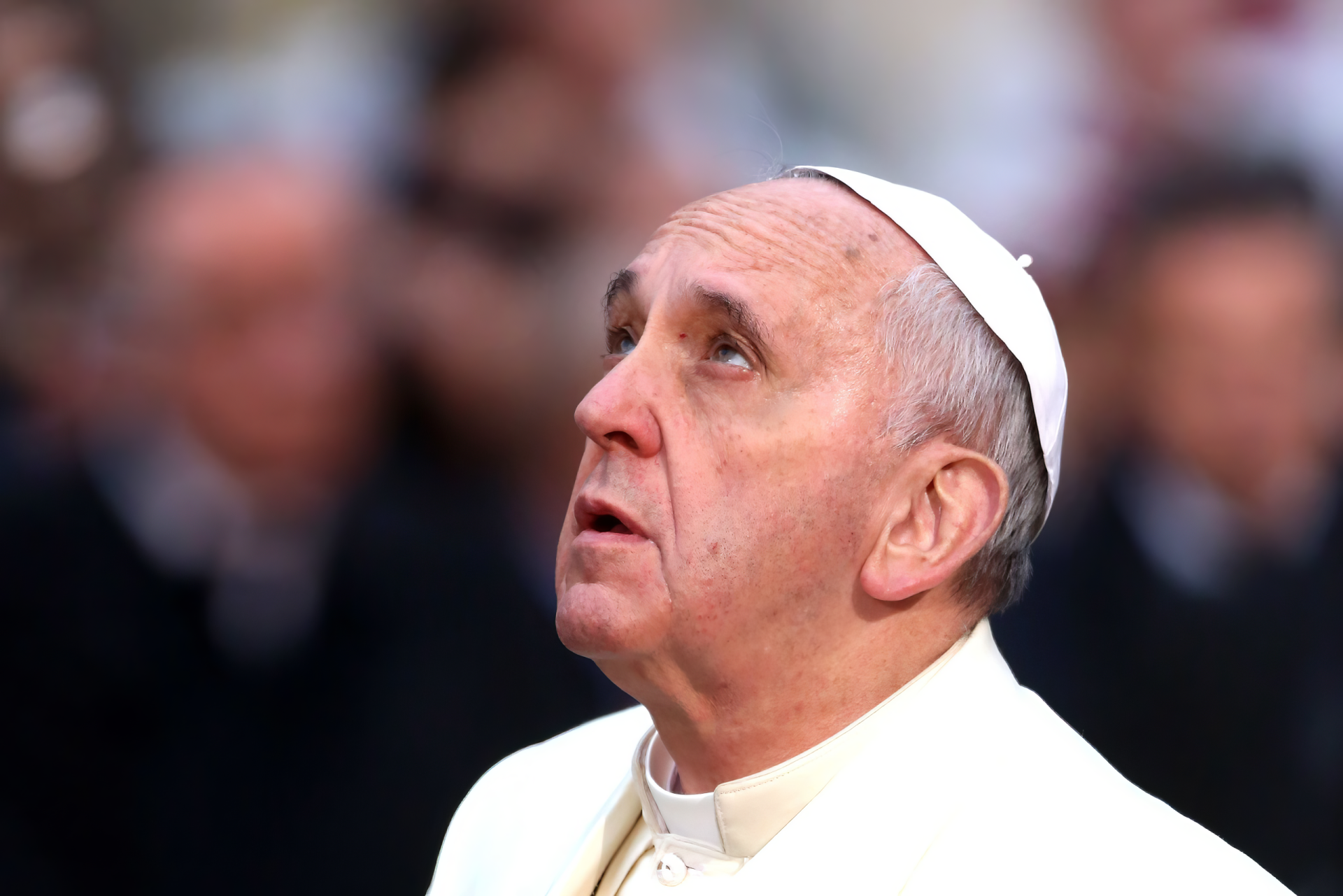 Pope Francis gazes upward, surrounded by a blurred background, capturing a moment of reflection. This HD image serves as a desktop wallpaper and background celebrating religious devotion.