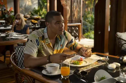 A scene from the TV show The White Lotus, featuring Nicholas Duvernay seated at a table, enjoying a meal, with a vibrant background that captures the show's tropical ambiance.